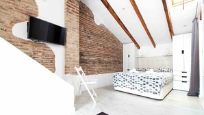 A bedroom with a bed, a TV mounted on a brick wall, a white chair, and a wooden ceiling beam. - Studio for couples with terrace on the beach (València)