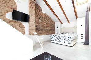 A bedroom with a bed, a TV mounted on a brick wall, a white chair, and a wooden ceiling beam. - Studio for couples with terrace on the beach (València)