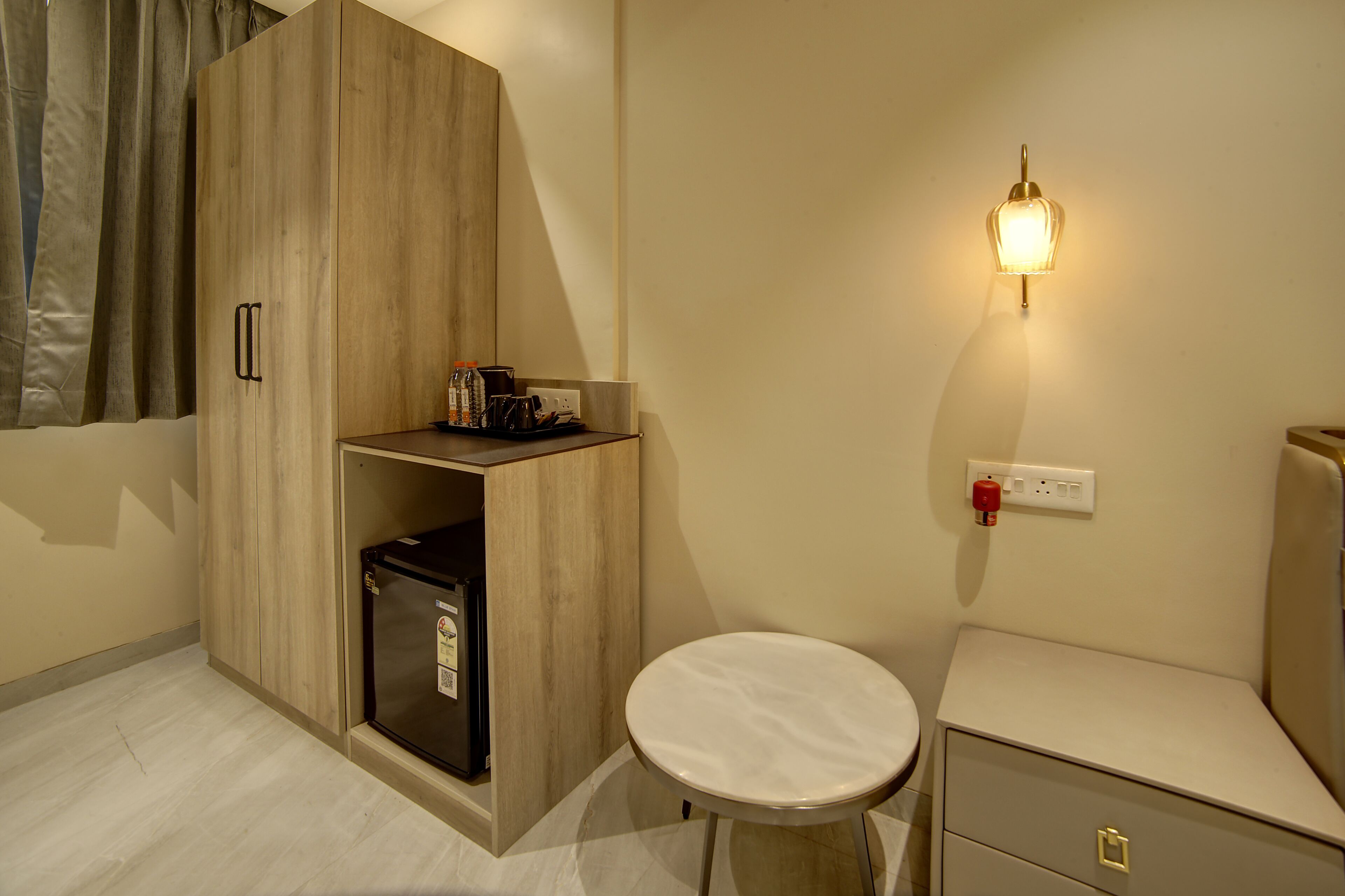A compact room with a wooden wardrobe, a small refrigerator, a round table, a bedside cabinet, and a wall-mounted light fixture.