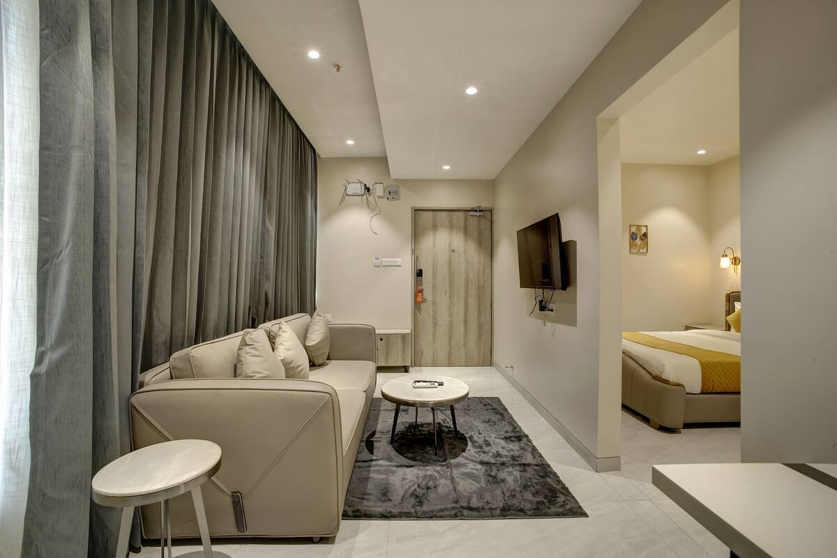 A modern hotel room with a sofa, a round coffee table, a bed with a yellow blanket, a TV mounted on the wall, and a bathroom visible through an open door.