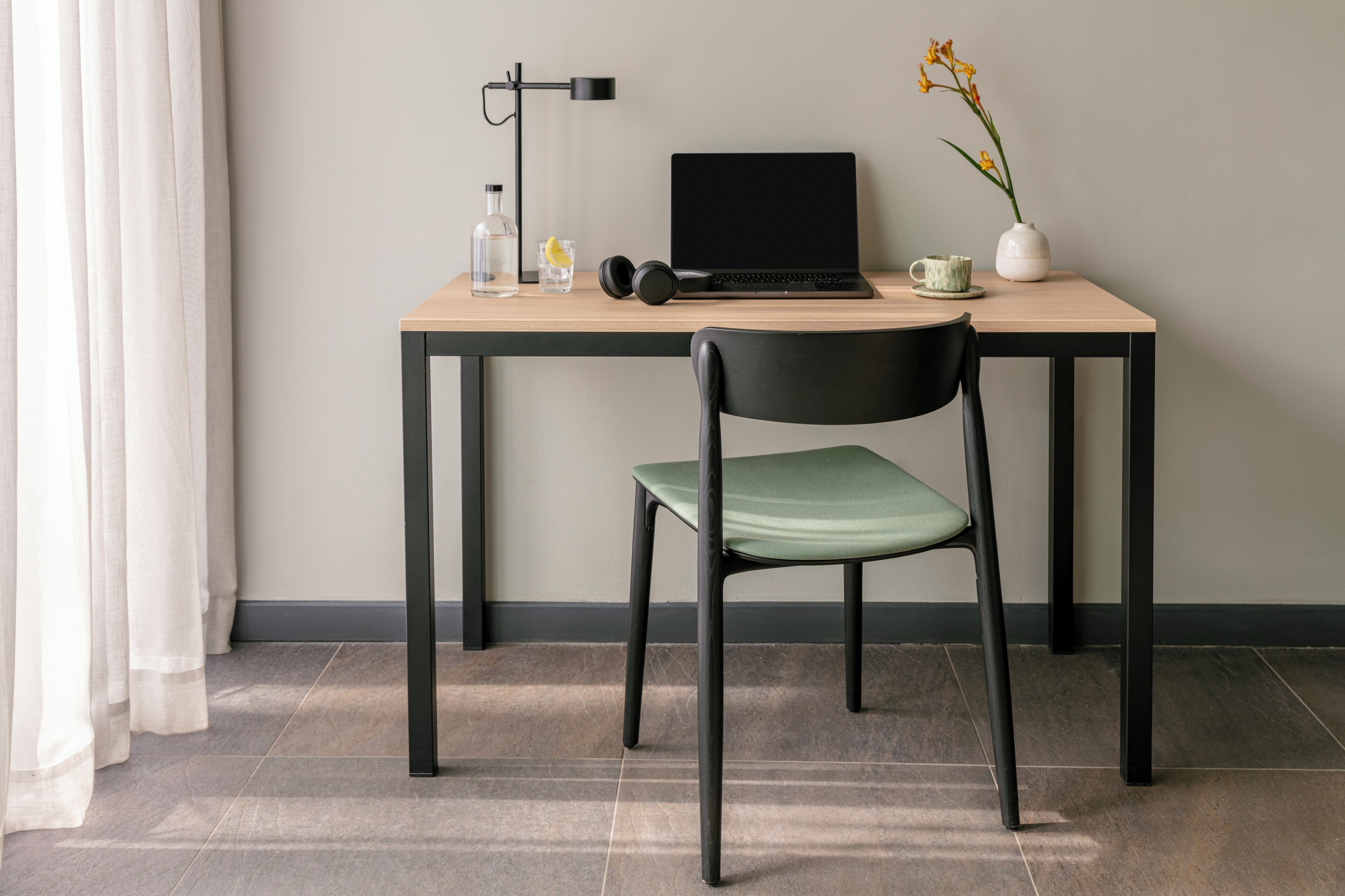 A workspace with a laptop, headphones, a bottle, a cup, and a vase with flowers on a wooden desk.