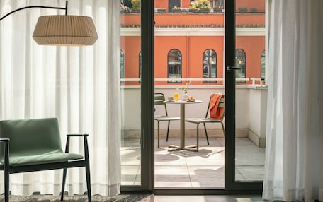 A room with a green chair, a table with a lamp, and a view of a building with red walls through a glass door.