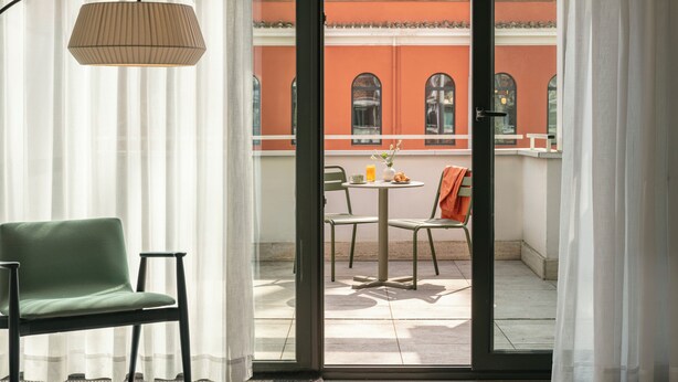 A room with a green chair, a table with a lamp, and a view of a building with red walls through a glass door. - Bob W Rome Testaccio (Rome)