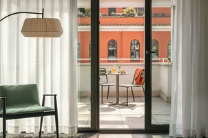 Ein Zimmer mit einem grünen Sessel, einem Tisch mit einer Lampe und Blick auf ein Gebäude mit roten Wänden durch eine Glastür.