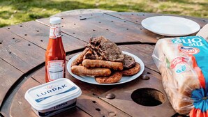 A picnic setup with a plate of food, a bottle of ketchup, a container of yogurt, and a package of bread on a wooden table.