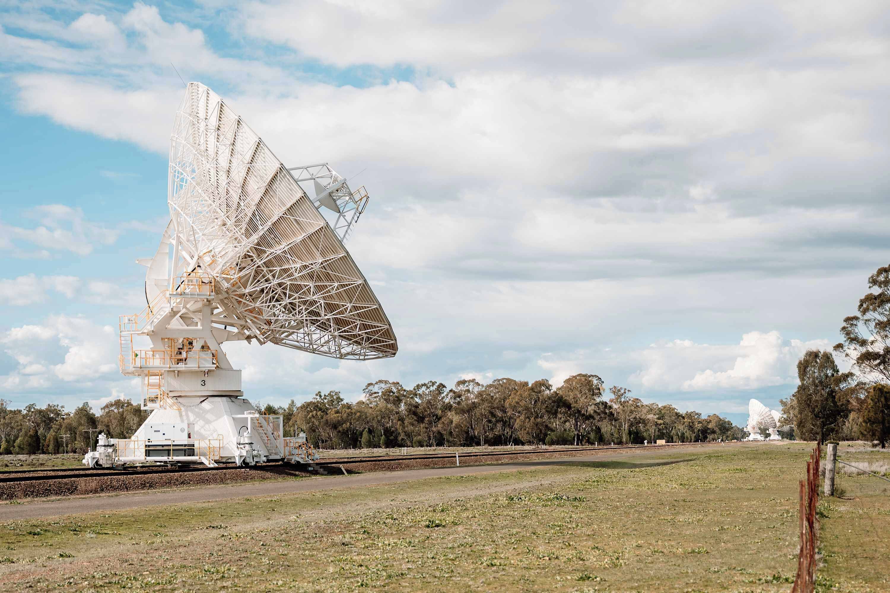 En stor, hvid satellitantenne i et åbent marklandskab med træer i baggrunden.