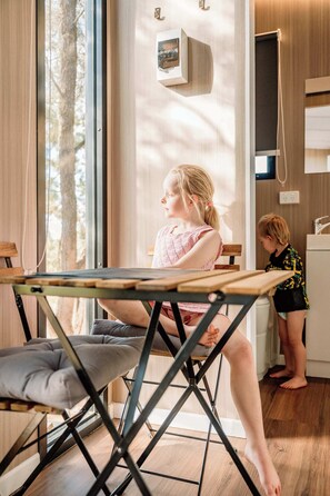 Una habitación con un ventanal grande, suelo de madera, una mesa con cojín y una niña planchando ropa.