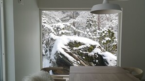 Un intérieur moderne avec une table à manger en bois, un chien blanc au pelage duveteux et une vue sur un paysage enneigé par une grande fenêtre.