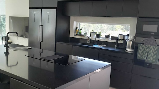 Une cuisine moderne dotée d’un îlot central, de meubles de rangement aux couleurs sombres et d’une fenêtre qui laisse entrer la lumière naturelle.