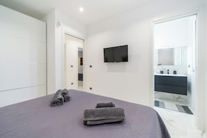 Une chambre à coucher comprenant un lit, un téléviseur à écran plat fixé au mur, une salle de bain avec un miroir et un lavabo, et une porte donnant sur une autre pièce.