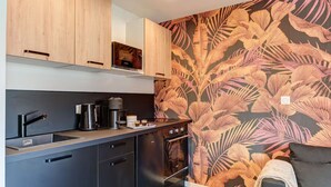 A compact kitchen with a modern design, featuring wooden cabinets, a black countertop, and a decorative leaf-patterned wall. - Lyon Est Élégance - Parking & Comfort (Meyzieu)