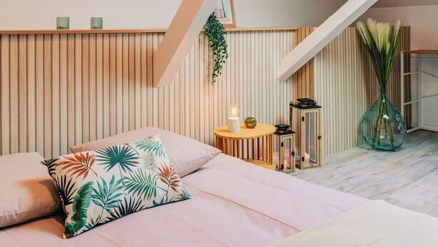 A bedroom with a sloped ceiling, a bed with floral pillows, a small round table with a candle, and a vase with plants. - Lyon Est Élégance - Parking & Comfort (Meyzieu)