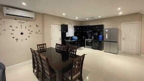 Een eetgedeelte met een tafel en stoelen, een keuken met zwarte kastjes, een koelkast en een airconditioner.