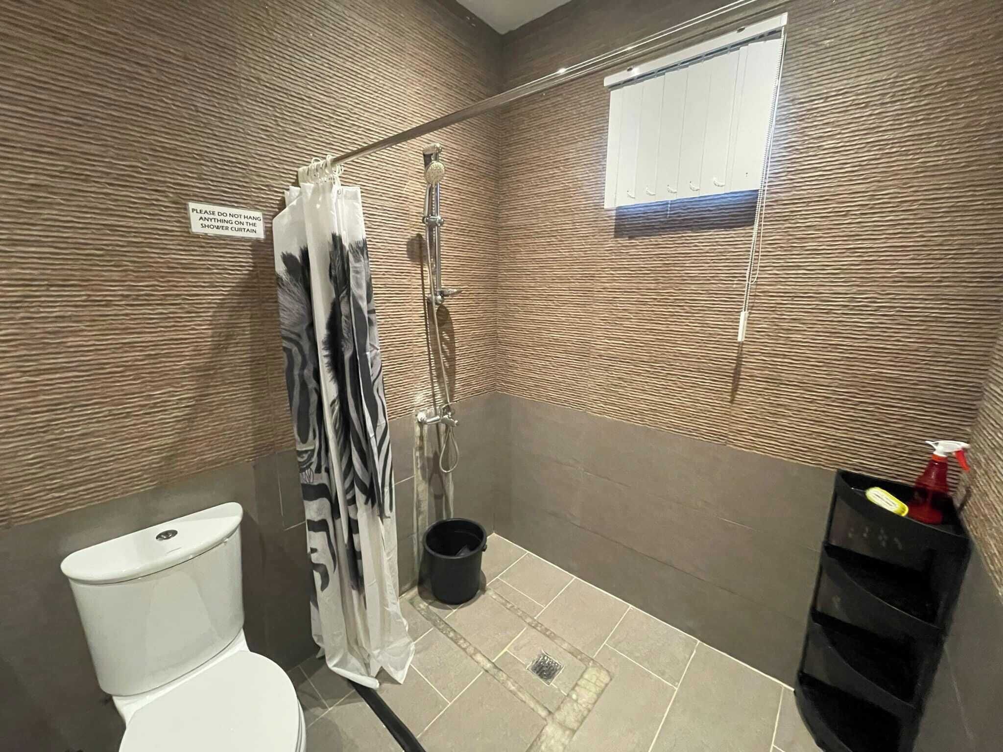 A bathroom with a shower curtain, a toilet, a bucket, and a shelf with cleaning supplies.