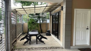 Een overdekt terras met een tafel en stoelen, een deur en uitzicht op een tuin met palmboompjes.