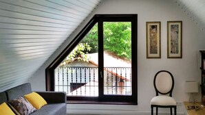 Ein Zimmer mit schrägen Wänden, einem grauen Sofa mit gelben Kissen, einem weißen Sessel und einem großen Fenster mit Blick auf Grünflächen und ein Gebäude.