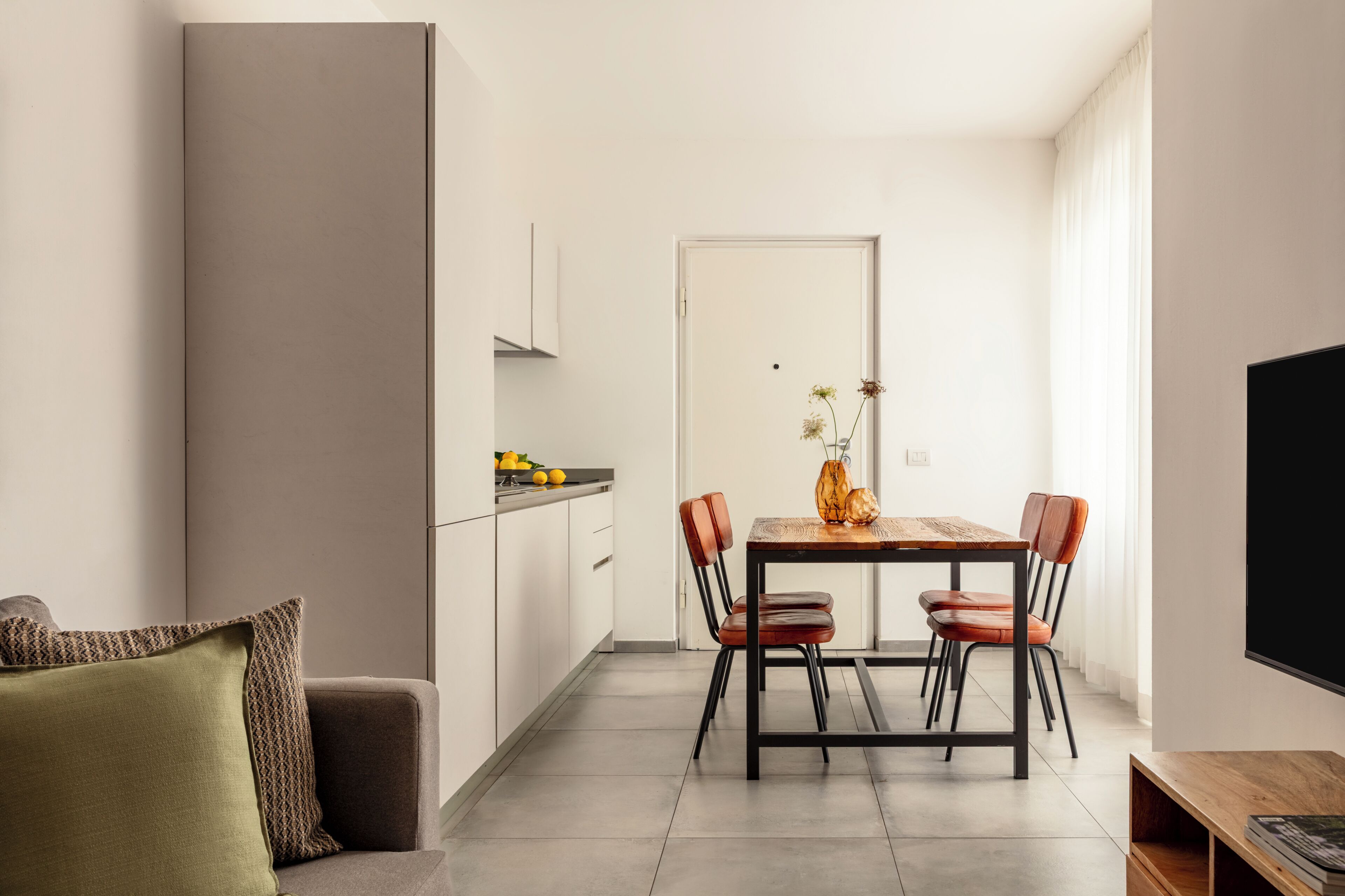 A modern kitchen with white cabinets, a dining table with chairs, a sofa with a patterned cushion, and a flat-screen TV mounted on the wall.