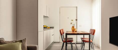 A modern kitchen with white cabinets, a dining table with chairs, a sofa with a patterned cushion, and a flat-screen TV mounted on the wall.