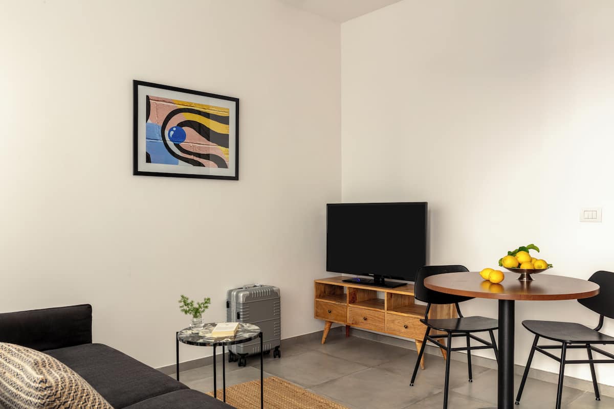 A modern living room with a sofa, a dining table with chairs, a television, a small side table, and a suitcase.