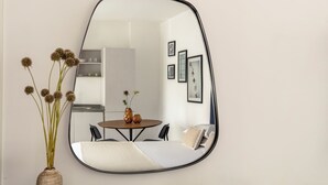 A mirror with a round table and chairs in front, a bed with pillows, and a vase with flowers to the left.