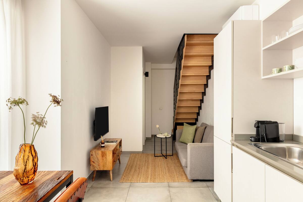 A modern living space with a sofa, coffee table, TV, wooden staircase, and kitchen area.