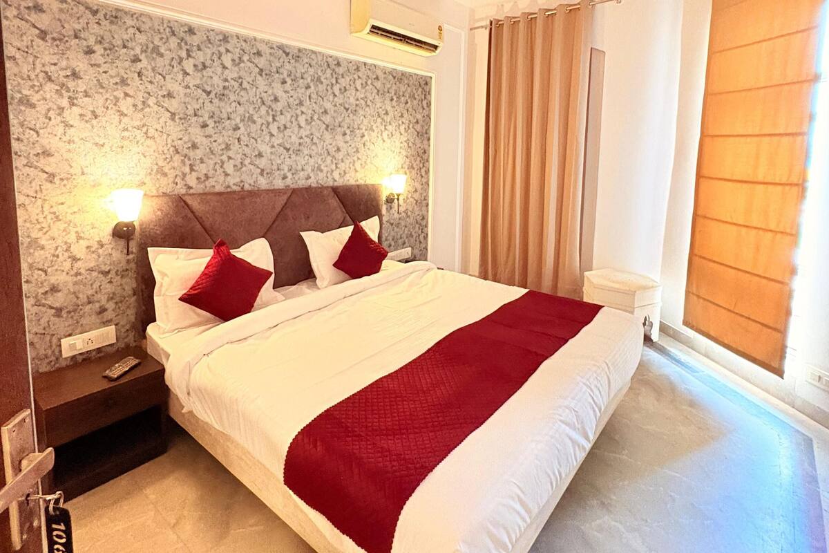 A hotel room with a bed, a ceiling fan, an air conditioning unit, a nightstand, a remote control, and a window with curtains.