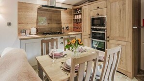 A modern kitchen with wooden cabinets, a built-in oven, and a dining area with a table set for two. - The Pines - Kelling Heath Holiday Park - 19233 (Holt)