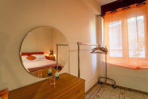 Une chambre avec un miroir rond, une commode en bois, une table de chevet avec un verre d’eau, un porte-vêtements suspendu et une fenêtre avec des rideaux.