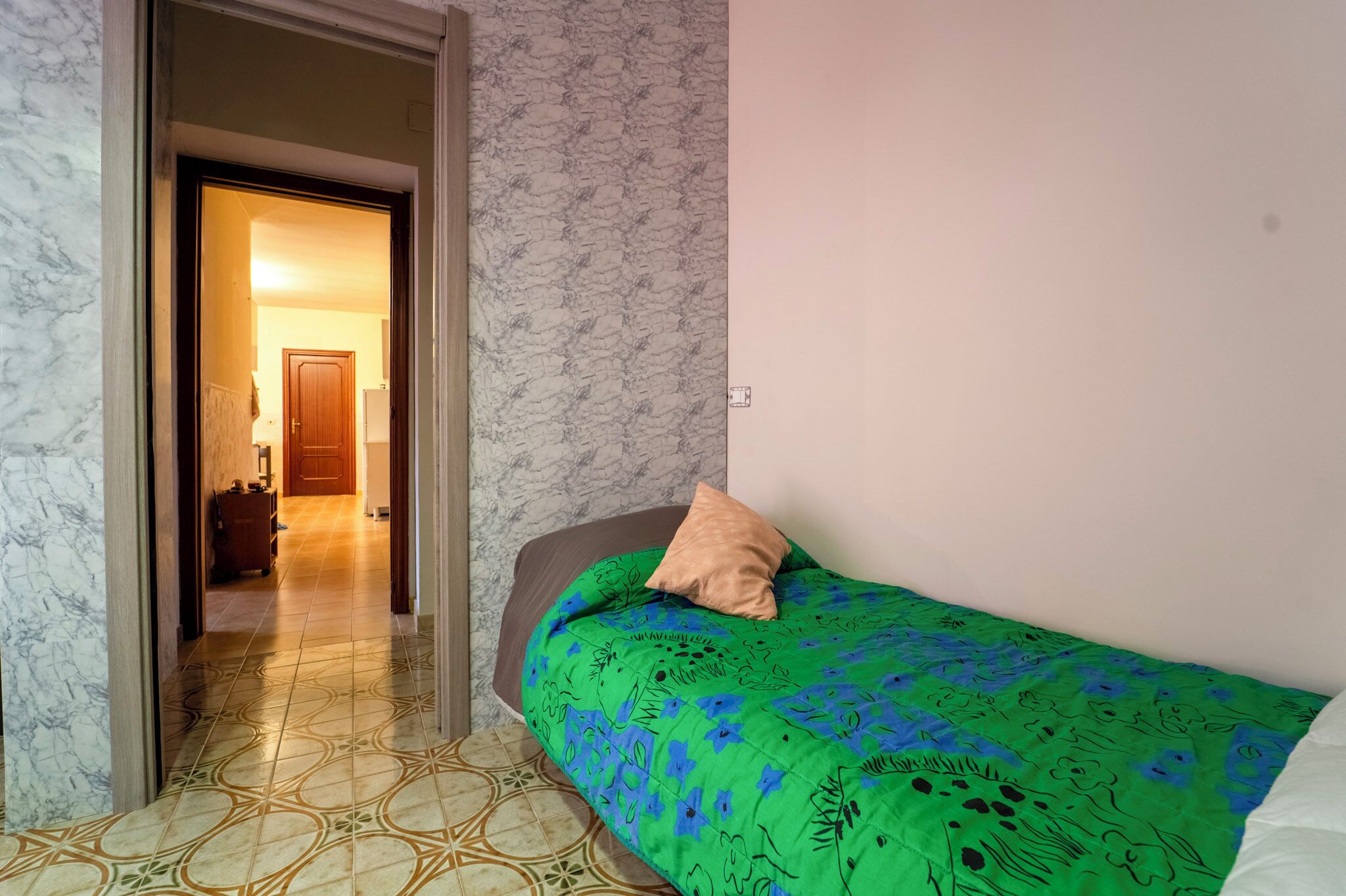 A bedroom with a bed, a door leading to another room, and patterned flooring.