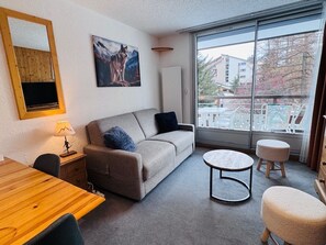 A modern living room with a sofa, a round coffee table, a wooden dining table, and a large window offering a view of buildings and trees. - Charming studio in the heart of Les Deux Alpes for 4 people (Les Deux Alpes)