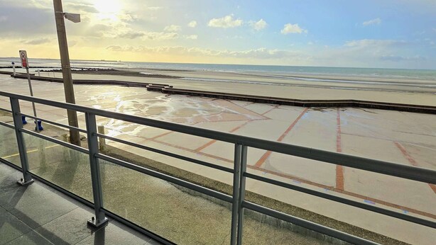 Blick auf einen Strand von einer überdachten Fläche mit Metallgeländer, bewölkter Himmel und ruhiger See.