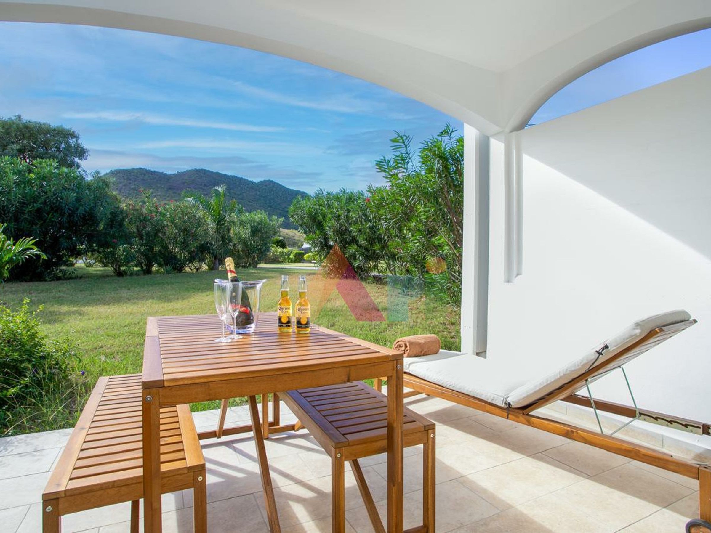 Una mesa de madera con dos sillas, una copa de champán, una botella de Corona y una toalla en una silla de descanso blanca, con vistas a un jardín exuberante y montañas.