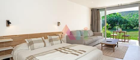 A modern bedroom with a large bed, a sofa, and a dining area with a view of greenery.
