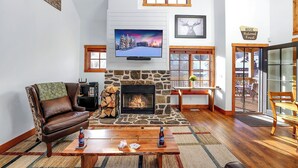 A cozy living room with a stone fireplace, a leather armchair, a wooden coffee table with drinks, and a television mounted above the fireplace. - Ski in/out to Tamarack Express Chairlift, pet-friendly, AC, hot tub (Tamarack)