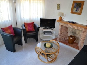 Una sala de estar con dos sillones de cuero negro, un televisor sobre un mueble blanco, una silla de mimbre con una vela y una chimenea de ladrillo.