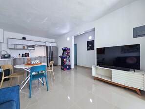 Ein modernes Wohnzimmer mit Flachbildfernseher, einem Essbereich mit Stühlen, einer Küche inklusive Kühlschrank und einer Spielkonsole.