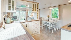 A modern kitchen with white cabinetry, a built-in oven, and a dining area with a table and chairs. - Two Hoots - Kelling Heath Holiday Park - 19668 (Holt)
