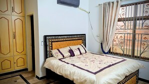Une chambre à coucher comprenant un lit, un appareil de climatisation, une fenêtre ornée de barreaux en fer décoratifs et une porte au design traditionnel.