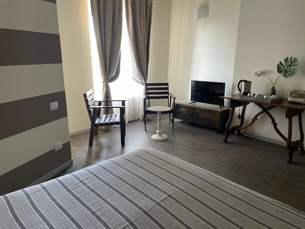 A hotel room with a bed, a TV, a desk with a coffee maker, a chair, and a plant.