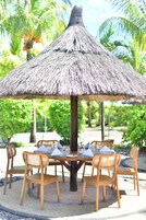 Una mesa con sombrilla de paja y seis sillas de madera, rodeada de follaje y palmeras.