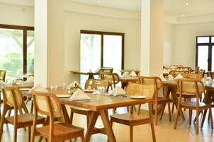 Un comedor con mesas y sillas de madera, distribuido en un espacio amplio con grandes ventanales que dejan entrar luz natural.