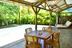 Una mesa de madera con cuatro sillas, una pared de piedra, una piscina y un área de descanso con un sofá blanco y una tumbona.