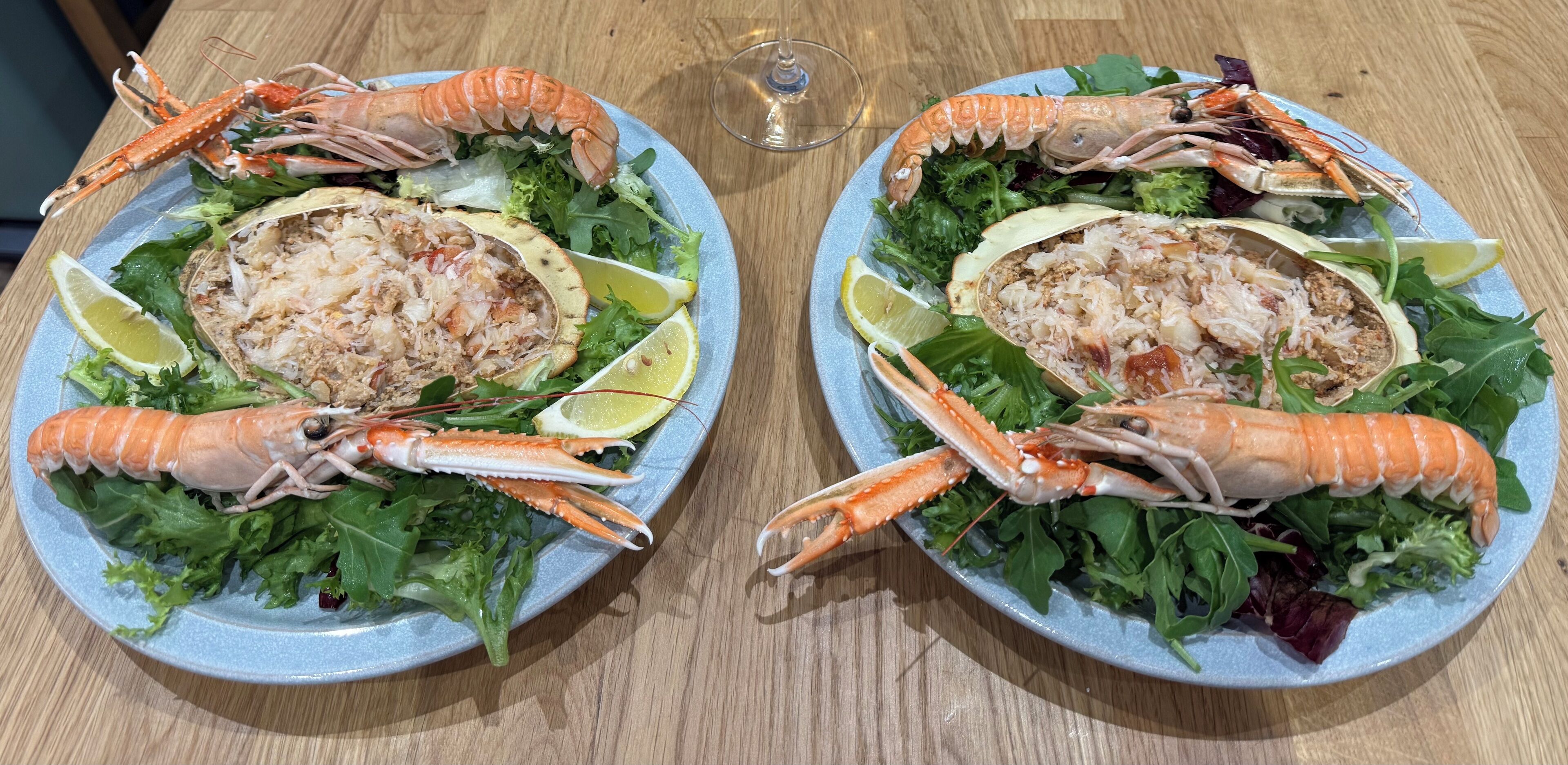 Two plates of seafood with lemon slices and greens on a wooden table.