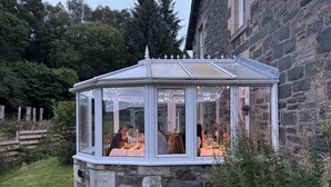 A glass-roofed gazebo with a stone base, surrounded by greenery and a stone building. - Crannaig House (Stirling)
