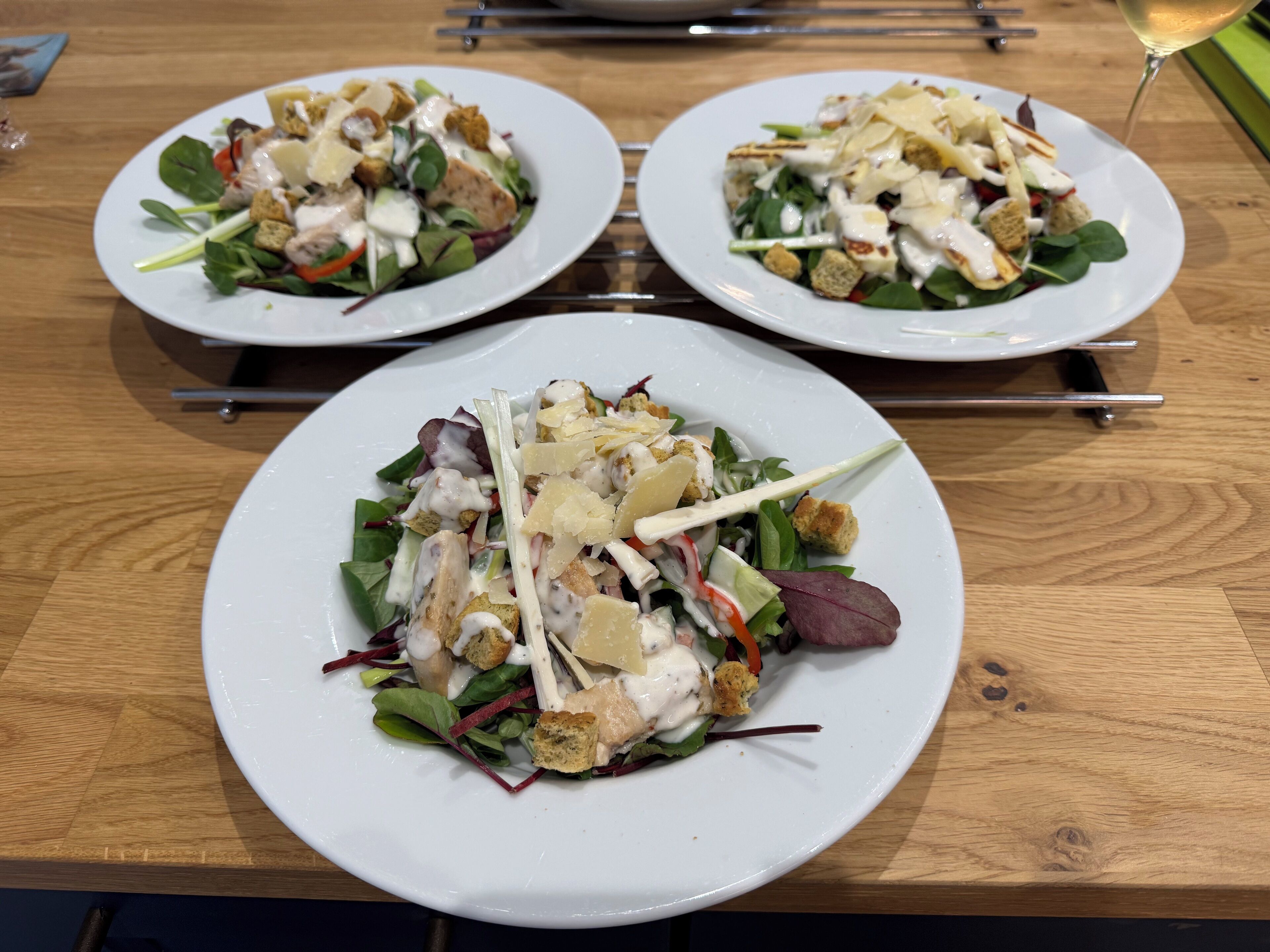 Drie witte borden met salade, croutons en kaas op een houten tafel.