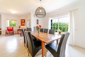 A dining area with a wooden table, black chairs, a hanging light fixture, and a window with a view of greenery. - Villa Nox is perfect for families with children as the pool is fenced (Albufeira)