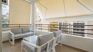 En balkon med sofa og stole, et glasbord og udsigt til bygninger.