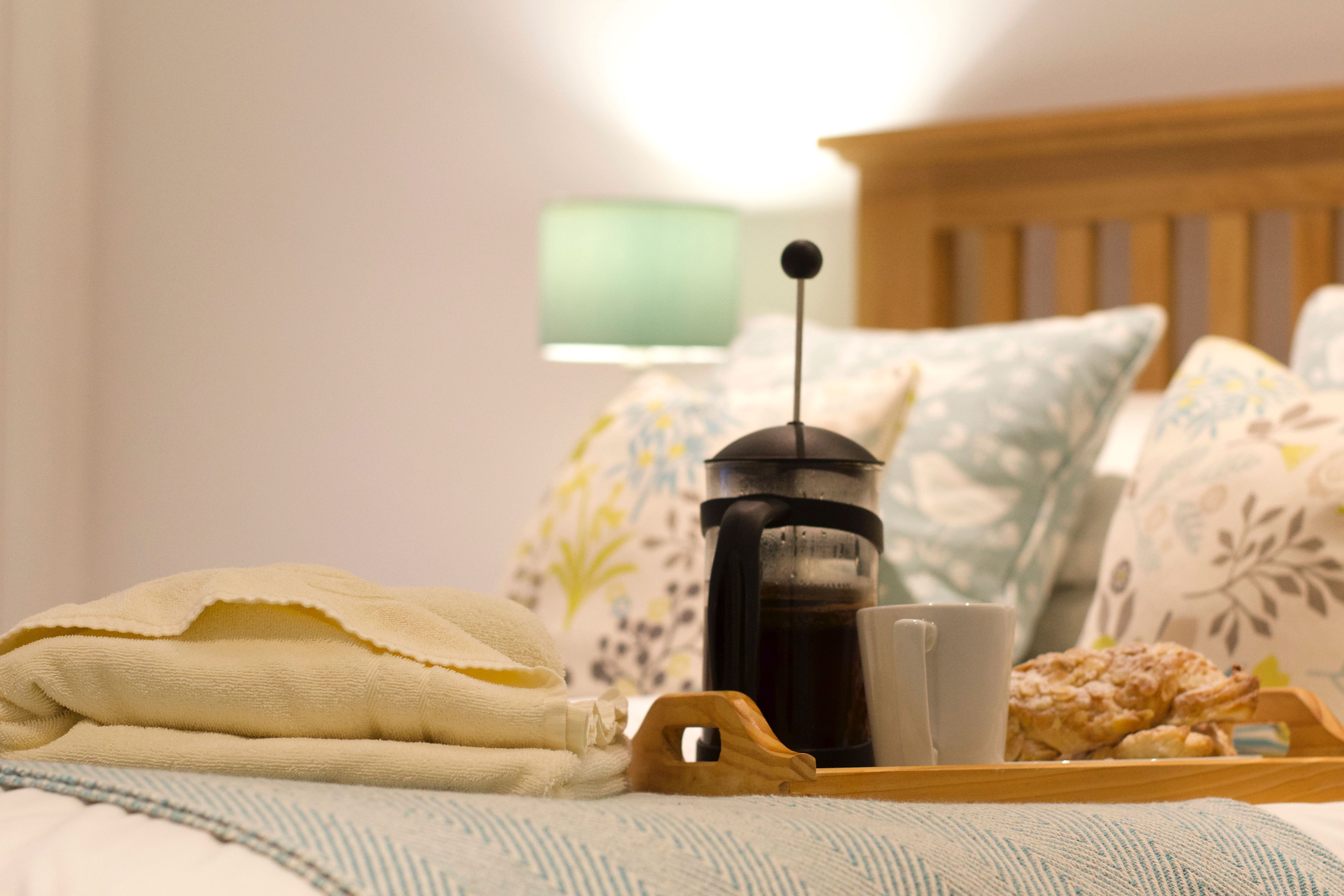A bed with a wooden headboard, a coffee pot, a cup, and a tray with a pastry.