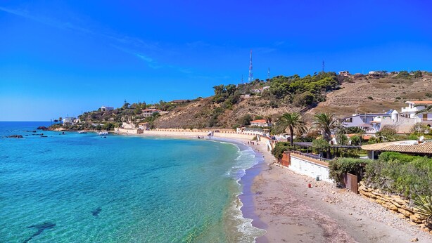Une plage côtière aux eaux d’un bleu limpide, avec un rivage sablonneux et des bâtiments construits sur une colline.