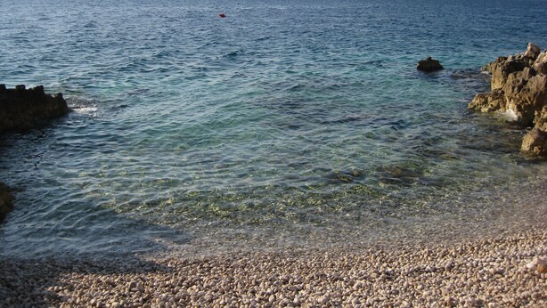 En strand med småsten, klart vatten och berg i fjärran.